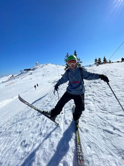 An individual is depicted on a snow-covered mountain slope in Jahorina, Bosnia and Herzegovina, under a clear blue sky. The person, positioned centrally, is a male wearing a bright green ski helmet, white-framed goggles with an orange tint, a blue denim-style half-zip jacket with "VOLCOM" lettered in red across the chest, black ski pants, and black gloves. He is equipped with skis and poles, with his left ski visibly lifted and his right ski planted on the snow. His arms are extended slightly, holding a black ski pole in each hand. A black backpack is worn on his back. His mouth is open, suggesting speech or an expression.
The foreground consists of disturbed snow, showing ski tracks and shadows. In the midground, the snow-covered slope extends upwards, revealing multiple ski lift towers and cables. Several distant figures are visible; two individuals are walking uphill on the left side of the frame, and another person is situated further right beneath a ski lift. Scattered evergreen trees are present on the slopes. The background features a prominent white, spherical domed structure, likely a radar station or observatory, atop a distant snow-covered peak. The sky is a uniform, clear blue, with a strong sun glare originating from the upper right portion of the image. The overall scene is indicative of a sunny winter day at a ski resort.
