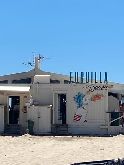 The image shows a beachfront building in Conil De La Frontera, Spain, with a clear blue sky overhead. The building is white with a light grey roof and features the words "UGUILLA Beach" in teal lettering above a stylized mural of a person with blue hair and a fish. To the left of the mural, a section of the building has a white awning over an entrance. A red fire extinguisher is mounted on the wall. A green trash can is positioned on the sand in front of a set of double doors. A satellite dish and antenna are mounted on the roof. The foreground consists of sand dunes.