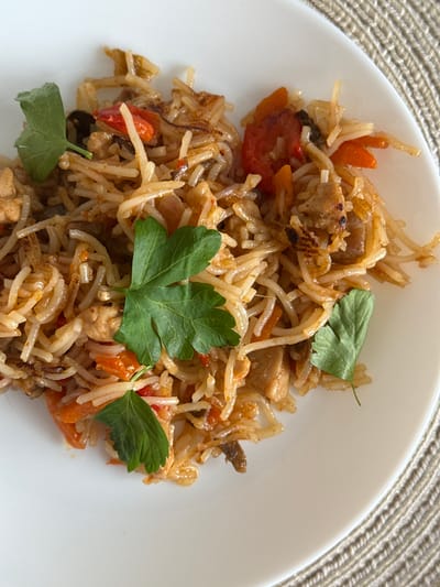 A close-up, overhead shot features a portion of a white plate filled with a portion of a rice dish. The rice appears to be mixed with small pieces of meat, carrots, and tomato, and is garnished with a few sprigs of fresh parsley. The dish is presented on a textured, beige placemat. The specific location in the city of Гамово, Russia, cannot be determined from the visual information provided.