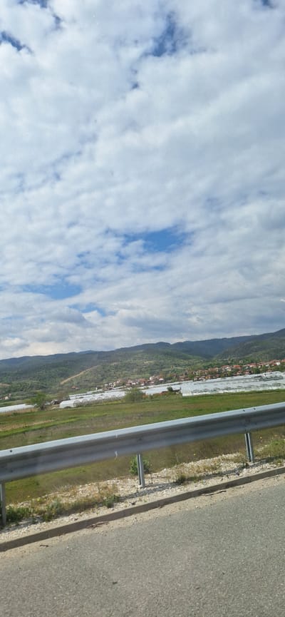 The image shows a view of Mikrevo, Bulgaria, from a roadside. The sky is filled with white, streaky clouds with patches of blue. A mountain range covered in green vegetation runs along the horizon. Near the base of the mountains, the town of Mikrevo is visible, with closely packed houses and structures. In the foreground, a guardrail runs parallel to a paved road. Beyond the guardrail, there is a strip of green grass. The perspective suggests the photograph was taken from a moving vehicle on the road.