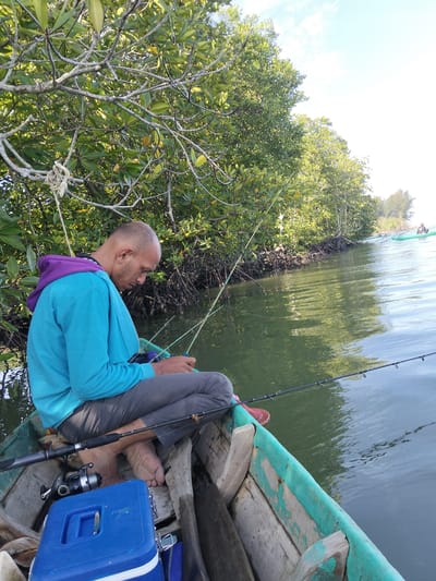 An adult male with a dark complexion, bald head, and facial stubble is seated barefoot in a small, weathered wooden boat in a waterway near Kualasimpangulim, Indonesia. He wears a teal-blue long-sleeved hoodie with a purple hood lining and dark grey trousers. The man is focused downwards, holding a fishing rod, with another rod visible resting on the boat's side, featuring a silver and black spinning reel. A blue plastic cooler with a white stripe is positioned in the foreground of the boat. The boat itself shows peeling light green/teal paint on its wooden gunwales.

The waterway features calm, dark green water. The left bank is densely lined with a mangrove forest, characterized by lush green foliage and visible aerial root systems extending into the water. A piece of white rope is tied from a tree branch in the upper left, suggesting the boat may be moored. In the distant background, another individual in a light green watercraft, possibly a kayak or small boat, is visible further down the waterway. The sky is bright and partly cloudy.