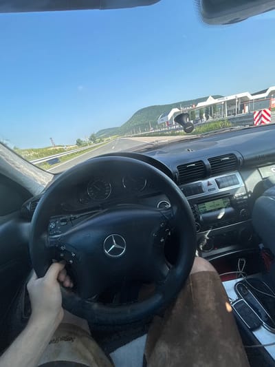 The image displays the interior of a Mercedes-Benz vehicle from the driver's perspective, looking forward onto a multi-lane highway in Mogila, Bulgaria. The sky is clear and bright blue.
The driver's left hand, adorned with a tattoo on the forearm, grips the black, three-spoke steering wheel featuring the Mercedes-Benz logo. The dashboard includes an instrument cluster with a speedometer calibrated up to 260 km/h and a tachometer. The center console contains air vents, a silver-colored panel with buttons (including ESP and hazard lights), and a car radio displaying "VESELINA" and "98.9 MHz". A black "Canyon" branded device is mounted on the dashboard. The driver's lap is visible, covered by brown shorts or pants. On the center console or passenger seat area, a smartphone, a car key fob, and charging cables are present. The passenger side dashboard displays an "SRS AIRBAG" label.
Through the windshield, a paved highway extends into the distance, bordered by a metal guardrail. Lush green hills and mountains are visible on the horizon. To the right of the road, a service area or gas station complex with "Vistran" and "power-X" branding is visible, fronted by a red and white striped barrier. On a distant hill to the left, a reddish-brown lattice tower stands prominently.