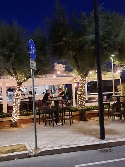 The image captures a nighttime scene on a street in Pesaro, Italy, likely near a waterfront promenade given the presence of trees and outdoor seating. A blue sign with a white bicycle symbol indicates a bike path. Beside it, a smaller rectangular sign reads "velocipedi a due ruote," confirming it's for two-wheeled vehicles.

In the background, a brightly lit establishment, possibly a cafe or restaurant, is visible. The building features large windows revealing a well-lit interior with what appears to be a counter and shelves stocked with bottles. The exterior of the building is adorned with string lights, and a tree in front of it is wrapped in numerous small, white lights, creating a festive ambiance.

Several tables and chairs are arranged on the sidewalk, some occupied. Two individuals are seated at a table on the left, facing each other, engaged in conversation. Another person is seated at a table on the right. The pavement is made of interlocking bricks, and a curb separates the sidewalk from the road, which is visible in the foreground and has a white line painted on it. A dark lamppost stands prominently in the right-center of the image. The sky is a deep, clear blue, characteristic of a night sky.