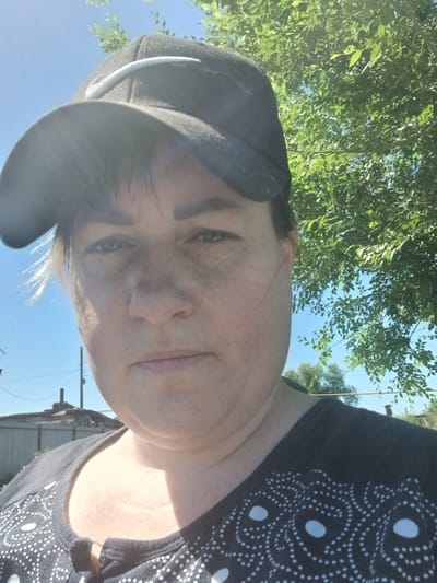 A woman wearing a black baseball cap and a black shirt with a white polka dot pattern is taking a selfie outdoors. She has light brown hair with some blonde highlights and a nose piercing. Behind her, a bright blue sky is visible with green tree leaves framing the top right corner. In the background, there are some structures that appear to be houses and a fence. The lighting suggests it is a sunny day.
