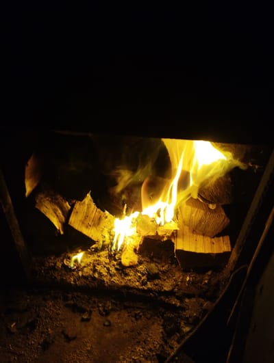 The image shows an indoor shot of a fireplace with burning logs. The fire is bright yellow and orange, with visible flames and smoke. Several logs are stacked within the fireplace, some of which are partially consumed by the fire. The background is dark, suggesting nighttime or an enclosed space. The scene appears to be a domestic setting, likely in a home or cabin. The location of Ширяево (ильинский), Russia, is not depicted in the image.