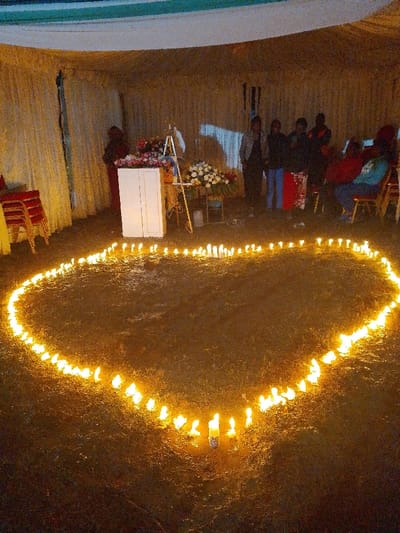 The image captures an outdoor gathering under a light-colored marquee tent on dark ground in Marenyo-Shianda ward, Kenya. In the foreground, a large heart shape is meticulously outlined by numerous lit white pillar candles. The background, dimly illuminated, reveals approximately 7-10 individuals. A cluster of 4-5 people, primarily women, stand observing in the center-right. Further right, 2-3 individuals are seated on red-backed chairs, one wearing a light blue hoodie. In the center-left, a white two-doored cabinet or podium is present, topped with a vibrant floral arrangement of red, pink, and purple flowers. To its immediate right stands a white easel and another floral arrangement of white and red blooms on a small stand. A stack of red chairs with gold frames is visible on the far left. The environment suggests a commemorative event or vigil.