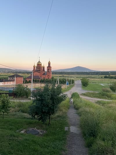 The image shows a scenic view in Novoterokskiy, Russia, likely at dusk. A prominent red brick church with golden domes stands in the mid-ground, a testament to local architecture and possibly religious significance. In the background, a large mountain looms under a clear, pale blue sky that transitions to a warm orange hue near the horizon, indicating the setting sun.
The foreground is dominated by lush green grass and trees, with a paved path winding through the landscape. A person is visible on the path, seemingly engaged in a run, adding a dynamic element to the tranquil scene. Further along the path, a small vehicle can be seen, suggesting the presence of a road or access route. The overall atmosphere appears peaceful and serene, with the soft light of dusk casting long shadows and highlighting the natural beauty of the surroundings. There is no visible text in the image.