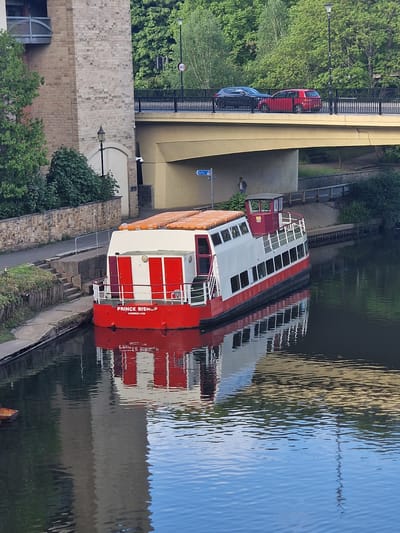 A boat, named "Prince Bishop Sunderland", is docked on a river in Durham, United Kingdom. The boat is white and red and has a glass enclosed top deck. It is docked against a stone and cement embankment. A sidewalk runs along the river bank. Above the boat is a bridge, with two cars driving on it. Trees and bushes are seen along the bank. The river water reflects the bridge and surrounding buildings.
