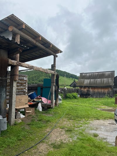 The image depicts an outdoor scene with a rustic, unfinished wooden structure in the foreground, resembling an open-sided shed or lean-to. The structure has a corrugated metal roof supported by rough-hewn wooden logs and poles. Construction materials such as lumber and concrete blocks are visible. To the left of the structure, a wall constructed from logs with visible chinking is present. Various objects are stored or piled within and around the structure, including a large blue barrel, cardboard boxes, tarps, and a green metal sheet. A black electrical cable runs across the ground from the foreground towards the right side of the frame.
In the background, a weathered wooden building with a corrugated roof stands to the right. Beyond this building, a wooden fence separates the yard from a grassy hillside covered in trees. The sky is overcast with gray clouds. The ground in the foreground is a mix of gravel and grass, with several puddles indicating recent rainfall. The overall impression is that of a rural or semi-rural setting, likely a homestead or a workshop area. The image appears to have been taken in Уршады, Russia, based on the provided location context. No people or animals are visible in the frame.