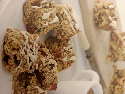 In Rumaithiya, Kuwait, a close-up shot showcases a cluster of homemade granola bars, artfully drizzled with white icing. These bars, packed with oats and dried fruit, are arranged on a white surface, possibly a plate or placemat. In the background, a portion of a white ceramic mug is visible, hinting at a cozy, domestic setting. The lighting is soft, creating a warm and inviting atmosphere, and the focus is sharp on the granola bars, emphasizing their texture and appealing appearance.