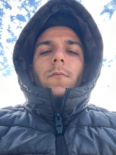 A low-angle selfie captures a young man wearing a dark, quilted jacket with the hood up. His face is tilted downward, showcasing dark eyes, eyebrows, and a faint mustache and goatee. The background reveals a cloudy sky. There is no indication that the photograph was taken in Srednia, Bulgaria.
