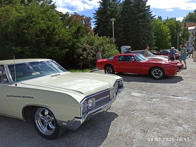 This is a full-frame, eye-level outdoor shot of a classic car show or gathering. In the foreground, a pale yellow classic American car, likely from the 1960s or 1970s, is prominently displayed, showcasing its chrome grille, headlights, and a shiny chrome rim on its front wheel. To the right and slightly behind it, a vibrant red classic American sports car, also from the 1970s, is parked. Several people are visible in the background, engaged in conversation or observing the vehicles. One man in a gray t-shirt stands near the red car, looking towards it. A woman in a blue dress and a man in a dark blue shirt are talking to each other next to the red car. Further back, other individuals and cars are present, and a portable toilet is visible. The scene is set against a backdrop of lush green trees and a bright blue sky with scattered clouds. The ground is paved with asphalt. A watermark in the lower right corner indicates the date "04 07 2026 16:17". While the precise location in Mariánské Lázně, Czechia, is not definitively identifiable from the image alone, the presence of classic American automobiles and the general ambiance suggest a public event or gathering.