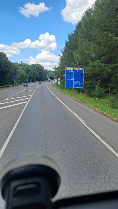 The image shows a view from a vehicle driving on a German autobahn. A large blue road sign is visible on the right side of the road, indicating directions to Siegen, Dillenburg, Dortmund, and Gießen. The autobahn has multiple lanes and is bordered by a dense forest on the right. On the left side of the autobahn, there are trees and a sound barrier. The sky is blue with white clouds. The foreground of the image is out of focus, showing a part of the vehicle's interior, possibly a dashboard mount. Based on the road sign showing "Siegen" and "Dillenburg", the location is on the Autobahn 45, likely in the vicinity of Burbach, Germany, as these are major cities and towns in the administrative region of Burbach.