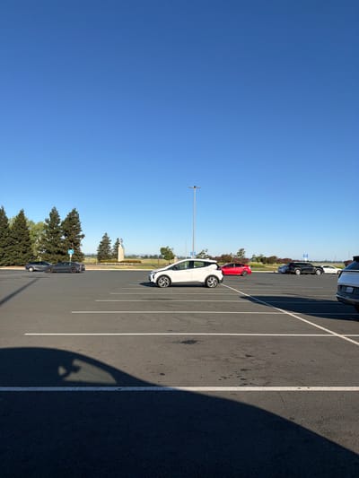 An outdoor, medium-wide shot captures a parking lot under a clear blue sky. In the foreground, a faint car shadow is cast across the asphalt, which is marked with white parking lines. A white electric car is parked in the middle ground, to the right of center. Further back, a red car is visible, followed by a black SUV and a white sedan. On the left side of the frame, two dark gray sedans are parked. In the background, a row of tall evergreen trees stands behind a low fence. Beyond the trees, a light-colored structure with blue accents rises, possibly a monument or building. The overall scene is clean and orderly, with no people visible. The image appears to have been taken in Sacramento, California.