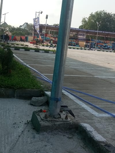 A metal pole stands on a concrete base with visible bolts and construction debris. Two blue hoses are loosely coiled near the base. A grassy area with small plants is to the left. In the background, a structure with signage related to a basketball championship is visible, with figures walking around. The area appears to be a parking lot or outdoor venue, with painted white lines marking parking spaces.