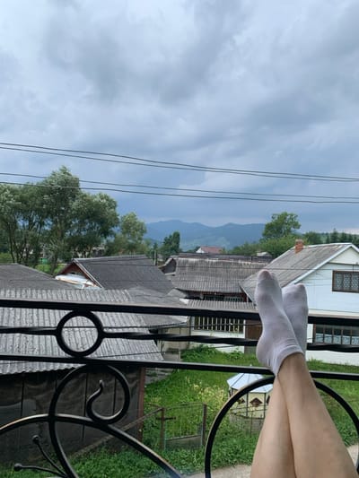 The image depicts a first-person view from a balcony overlooking a residential area in Lazeshchyna, Ukraine, with a mountainous backdrop under an overcast sky. In the foreground, the lower legs and feet of an individual wearing white ankle socks are elevated and crossed, resting on or over a black wrought-iron balcony railing with decorative circular and scrolled elements. The railing occupies the lower portion of the frame. Below the railing, several houses with grey corrugated roofs are visible. To the immediate right, a white-walled house with brown window frames and a red-tiled chimney is prominent. Green foliage, grass, and a metal gate are visible between the structures. In the midground, various trees with light green leaves are present, and three black utility lines traverse horizontally across the upper mid-section of the image. In the background, dark blue-grey mountains or hills rise under a uniformly cloudy, light grey sky. A small, light-colored gazebo is visible below the person's feet.