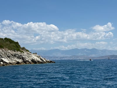 A wide shot captures the azure waters of the Ionian Sea, with a rugged, green-topped rocky coastline on the left. The sea is a deep blue, with gentle waves creating a textured surface. In the distance, hazy blue mountains form the backdrop, suggesting a coastal mountainous region. The sky above is a bright blue, dotted with fluffy white cumulus clouds. A speed boat is visible in the middle right of the frame, leaving a white wake as it cruises across the water. Further to the right, a tall, dark structure resembling a buoy or marker stands in the sea, with a smaller structure next to it. The scene suggests a serene summer day in a Mediterranean coastal area, likely within Kassopaia Municipal Unit, Greece, known for its scenic coastline and proximity to mountainous terrain.