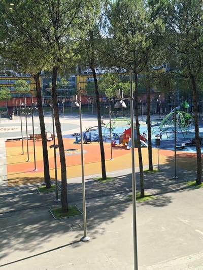 The image depicts an outdoor playground area in a city. In the foreground, several tall, slender trees are prominent, casting shadows on a paved ground surface. The playground itself features a colorful, multi-level structure with slides, climbing nets, and swings. The ground surface of the playground is a mosaic of vibrant colors, including orange, yellow, and blue, suggesting it is designed for children. In the background, modern buildings with large windows are visible, indicating an urban setting. The overall scene is bright and appears to be a sunny day. The location of this playground in Nola, Italy is not discernible from the image itself.