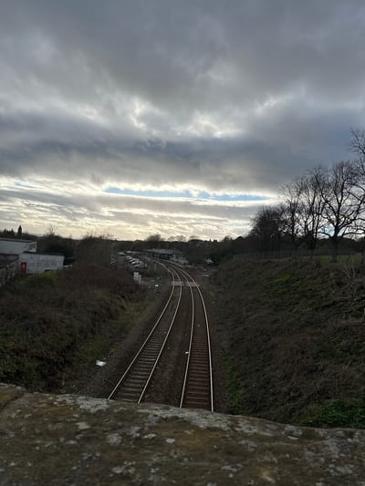 An elevated view overlooking railway tracks in Dunfermline, United Kingdom. The tracks curve through a landscape of dense shrubs and trees, adjacent to what appears to be a residential or commercial area with buildings and parked cars visible. The sky is overcast with layers of dramatic, textured clouds. The image appears to be taken from a stone bridge or retaining wall.