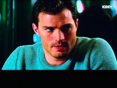 The image is a close-up, eye-level shot of a man's face and upper body, focusing on his head and shoulders. The man has dark hair, a light complexion, and is wearing a teal, cable-knit sweater. His left eye is prominently visible, appearing somewhat cloudy or reflective. His lips are a reddish hue, and he has stubble on his chin. In the upper right corner of the image, a logo reading "KINONOVA" is visible. The background is dark and out of focus, with indistinct colored lights, suggesting an indoor setting, possibly a room with ambient lighting or a screen. There is no identifiable connection to the city of Topolovgrad, Bulgaria.