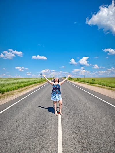 A woman stands with her arms raised on a deserted asphalt road under a bright blue sky with scattered white clouds. She is wearing a dark blue floral print tank top and a light blue denim skirt. The road stretches into the distance, bordered by a green field on the left and a grassy verge with some sparse vegetation and utility poles on the right. The woman's shadow is cast on the road. The scene is consistent with a rural or highway location outside of urban development. There is no indication of the specific location being in the city of Курово, Russia.