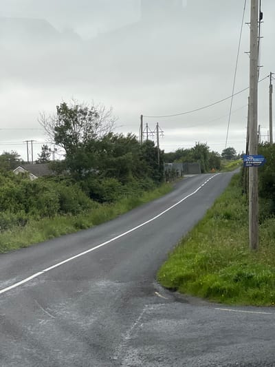 The image depicts a rural road curving to the right in Gweedore, Ireland. The road is paved and has a white line marking its center. On the right side of the road, a wooden utility pole stands tall, with a blue sign attached to it. The sign has white text that reads "CLUB EUROPA & CAMPERS" and "RYELEA". The surrounding environment is lush and green, with grass and trees lining the roadside. In the background, more trees and a house are visible, partially obscured by foliage. The sky is overcast and grey, suggesting a cloudy day. The scene appears to be empty of any people or vehicles, giving a sense of quiet solitude.