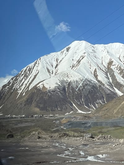 The image shows a mountain partially covered in snow, set against a blue sky. There are power lines visible in the upper left corner of the frame. The mountain appears steep, with visible rock formations and snow patches. The location is not specified.