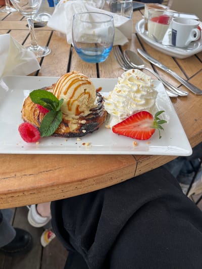 A white rectangular plate displaying a dessert is centrally positioned on a wooden outdoor dining table in Théoule-sur-Mer, France. The dessert features a round, partially charred, caramelized pastry topped with a scoop of vanilla ice cream drizzled with caramel sauce and sprinkled with crushed nuts. Adjacent to the ice cream, a sprig of mint and two raspberries are present. To the right, a dollop of whipped cream, also topped with crushed nuts, accompanies a halved strawberry with its green stem. The surrounding table surface includes an empty stemmed wine glass, a blue-tinted drinking glass, a crumpled white paper napkin, a fork, a knife, and a spoon. Further back, a white rectangular tray holds a small white espresso cup, a sugar packet, and two small glasses containing layered panna cotta-style desserts. In the lower foreground, parts of a person's upper thighs, clad in dark trousers, are visible. Below the table, portions of two pairs of feet rest on a wooden deck: one foot wears a black boot, and the other a white sneaker with yellow and red accents. The scene depicts a static meal presentation in an outdoor dining environment.