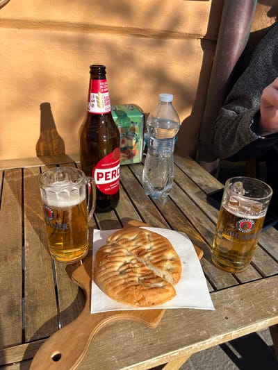 An outdoor scene in Monterosso al Mare, Italy, depicts a wooden slatted table illuminated by direct sunlight. On the table, from left to right, are a glass stein approximately half-filled with golden Paulaner beer and a foam head, an empty brown Peroni beer bottle with red labeling, a partially full plastic water bottle with a light blue cap, and a second glass stein approximately two-thirds full of Paulaner beer with minimal foam. A light wooden serving paddle holds a white paper napkin topped with a single piece of split focaccia bread. A green box with a fruit motif is visible behind the beer bottle and water bottle. A dark smartphone is partially visible on the table's right edge, adjacent to the right beer stein. The background features an orange-tan wall with distinct shadows cast by items on the table, and a dark grey vertical pipe is visible on the right side of the wall. The upper right quadrant of a person, wearing a grey speckled long-sleeved garment, is partially visible, with a hand also partly visible on the table.