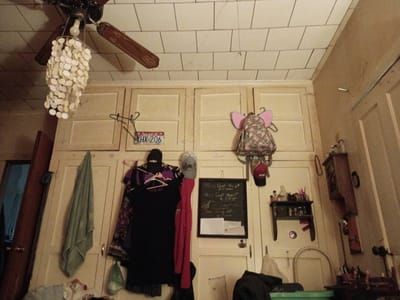 An eye-level indoor shot captures a cluttered room, looking towards a wall adorned with various personal items and a section of the ceiling. The ceiling features square light-colored tiles, from which a dark wooden ceiling fan with a decorative, shell-like chandelier hangs in the upper left.
The main wall beneath the ceiling is covered by light-colored wooden cabinets or built-in storage. Numerous objects are suspended from these cabinets and adjacent wall space. On the left, an Arkansas license plate displaying "XHX 206" is attached to a cabinet door. Several items of clothing, including a green towel, dark dresses, and patterned garments, hang on hooks and hangers. Two caps, one black and one grey/white, are visible above the clothes.
In the center, a small black chalkboard with white writing is hung, partially legible as "My Count My", "@ 9:30 Am" (or 9:50 AM), "The Covet Juve", and "Sam may 28". The word "Laugh" is faintly visible above the chalkboard. To the right, a floral backpack with distinctive pink "elephant ear" details hangs, alongside a red baseball cap. Further right, a small wooden shelf holds an assortment of bottles and small containers, while another, taller wooden structure with tools or implements is mounted on the wall.
A dark wooden doorway leading to a dim interior is visible on the far left. The bottom of the image shows glimpses of other items, including a green tub. The scene depicts a lived-in, domestic space in Clarksville, United States, characterized by its practical storage solutions and personal decorations. No direct activity is occurring, and the lighting suggests an indoor setting.