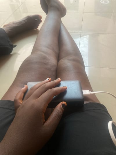 This media file captures a close-up view of a person's legs and hands, set against a tiled floor. The person is wearing a black top and appears to be holding a dark-colored power bank with a white charging cable connected to it. The person's nails are painted with a distinct design, featuring orange and black colors. Visible on the power bank is some faint text, though it is largely illegible. The lighting suggests it is daytime, and the setting is likely indoors, given the tiled flooring. The image is framed to focus on the person's legs and the power bank, with one foot also visible. The overall scene is casual and everyday.