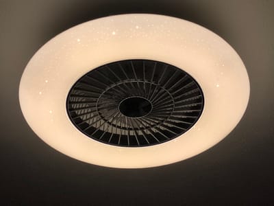 A flush-mount ceiling light fixture with an integrated fan is photographed from below. The fixture is circular and has a white outer ring with small, speckled light effects, resembling stars. The central part of the fixture contains a black fan with multiple blades, covered by a metal grille. The ceiling around the fixture appears to be a dark, textured surface. The image was taken in Dillingen an der Donau, Germany.