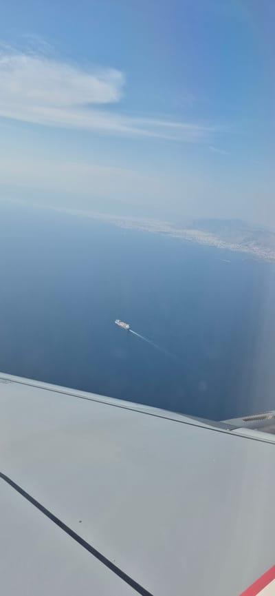 An aerial view captures the vast blue expanse of the Aegean Sea, with a hazy coastline of Spata-Loutsa Municipal Unit, Greece, visible in the distance. A cargo ship, painted in vibrant red and white, sails through the tranquil waters, leaving a white wake behind it. The foreground is dominated by the wing of an aircraft, its white surface reflecting the bright sky. Wisps of white clouds drift across the clear blue sky, hinting at the vastness of the atmosphere.