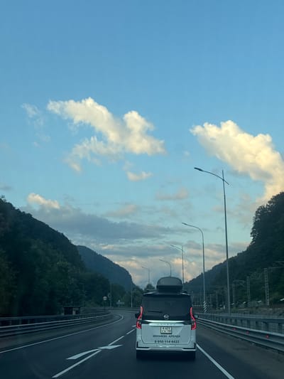 The image captures a scenic view from a moving vehicle on a highway in Chvizhepse, Russia. The road curves through a mountainous landscape with lush green slopes covered in trees. The sky is a mix of blue with scattered white clouds. Several streetlights line the highway. In the foreground, a white minivan with a roof carrier is visible, driving in the same direction as the camera. The minivan has a license plate and text in Russian on its rear. The road is paved with clear lane markings, and a metal guardrail runs along the side. The overall scene depicts a journey through a beautiful, natural environment on a well-maintained road.