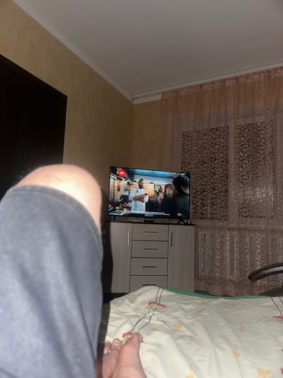 A person's left leg and foot are prominently featured in the foreground, obscuring much of the view. The leg is covered by dark grey shorts or trousers. The foot, appearing to be bare, is positioned at the bottom right of the frame, showing toes. In the background, a television is on, displaying a cooking show. A man in a chef's uniform is visible on the screen, with other individuals and a kitchen setting behind him. The television is placed on a light grey cabinet with drawers. To the right of the television, sheer pink curtains with a patterned design are drawn across a window. The room appears to be a bedroom, with a bed covered in a white and yellow patterned comforter visible in the lower right portion of the image. The walls are a muted yellow-brown color, and the ceiling is white with molding. The lighting suggests it is evening or indoors. The image does not contain any discernible elements that would place it specifically in Tselina, Russia.
