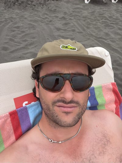 A man lies on a beach chair near black sand. He wears sunglasses and a khaki baseball cap with a yellow emblem. A colorful striped towel sits under him. A silver chain necklace is visible on his chest. He has a beard and mustache. The photo is taken from above, rotating the person 90 degrees counter-clockwise.