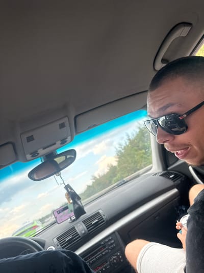 This image captures the interior of a vehicle's front cabin, providing an outward view of a roadside environment under a partly cloudy sky in Krushovitsa, Bulgaria.

A male individual is prominently featured on the right side of the frame, visible from the chest upwards. He has a very short, dark haircut and short facial stubble. He is wearing dark, reflective aviator-style sunglasses with a red and green stripe and a visible 'Gucci' logo on the right temple. His mouth is open, revealing his teeth. He wears a black short-sleeved garment and light-colored, possibly white or grey, shorts. His right hand holds a lit cigarette that emits smoke, positioned directly above a metallic silver can or container.

The steering wheel of the vehicle is partially visible on the lower left. The legs of a second individual, dressed in black fabric, are visible near the steering wheel.

The car's interior components visible include a gray fabric headliner, an overhead console containing lights and vents, and a rearview mirror from which a black air freshener dangles. A smartphone in a purple protective case is mounted on the dashboard, to the right of the rearview mirror. The dashboard is dark, featuring multiple air vents, a silver trim line, and a central console with various control buttons and displays.

Through the front windshield, a broad light blue tint band is present at the top. Beyond the windshield, a clear blue sky with white clouds is visible, along with a dense line of green trees and foliage suggesting a rural or suburban roadside. In the distance, a bright green vehicle is partially visible on the road ahead.