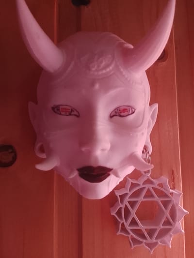 A white, horned mask with red and black makeup around the eyes and dark red lips is hanging on a wooden wall. A white, geometric star-shaped ornament hangs from a hoop earring on the mask. The photograph appears to have a reddish tint. The location is Высокиничи, Russia.