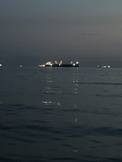 The image depicts a serene seascape at dusk, with a large cargo ship illuminated by its deck lights on the horizon. The ship, appearing to be a tanker, is positioned centrally, its lights reflecting on the calm water. Smaller lights, possibly from other vessels or buoys, are scattered across the water. The sky is a deep, dusky blue, transitioning to a darker shade above. The water surface is gently rippling, with subtle reflections of the lights. The scene evokes a sense of peacefulness and the vastness of the sea as day turns into night.