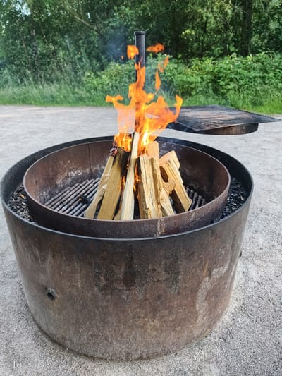 In Slaka, Sweden, a metal fire pit is centrally located on a gravel surface, surrounded by a wooded area. The fire pit, made of weathered steel, contains a burning wood fire, with flames reaching upwards. Inside the fire pit, a metal grill grate is visible, and a separate, larger grill grate rests on the edge of the fire pit. The background consists of dense green trees and vegetation, indicating an outdoor setting. The overall scene depicts preparation for or enjoyment of an outdoor fire.