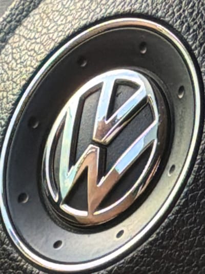 The image shows a close-up of a Volkswagen emblem, likely on a steering wheel or a car's exterior. The emblem is silver and chrome, with the iconic 'VW' design. It is set within a dark circular casing, which appears to be made of plastic or leather, with a textured surface surrounding it. There are several small, circular indentations or holes around the edge of the casing. The lighting in the image creates reflections on the metallic surface of the emblem. There is no indication of the city of Utrennyaya Dolina, Russia, or any other specific location in the image.