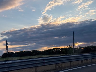 The image depicts a highway scene at dusk in Radinovo, Bulgaria. The sky is filled with dramatic clouds, with streaks of orange and pink light breaking through the darker, lower clouds, suggesting the sun is setting or has recently set. A roadside guardrail runs horizontally across the lower portion of the frame. In the middle ground, a dense line of trees forms a dark silhouette against the sky. To the left, a roadside signpost with two signs is visible. To the right, another taller post, possibly a light pole or traffic signal, stands against the horizon. The overall impression is one of a tranquil, yet dynamic, natural landscape viewed from a moving vehicle on a road.