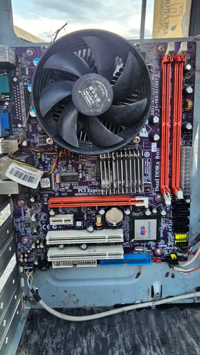 This image shows a close-up, overhead view of an open computer motherboard with its CPU cooler fan prominently displayed. The motherboard, which is purple with various components, appears to be situated within a computer case, suggesting it's part of a desktop computer. The scene is outdoors, indicated by the bright sky and what looks like a blurry background of clouds and possibly structures. The lighting suggests it might be daytime. There are no people visible in the image.
The motherboard itself is the primary subject, with visible text on it including "Dual DDR2 800," "PCI Express," "ELITEGROUP," and "THERMAL MASTER." There is also Chinese text which translates to "Cool Master." The motherboard has several slots for RAM, expansion cards, and various chips and capacitors. The CPU cooler fan is large and black, with "THERMAL MASTER" printed on its center. Cables are connected to some components.
The overall impression is that of a disassembled computer component, possibly being examined or prepared for repair or recycling, given the outdoor setting and the open nature of the computer case. The setting appears somewhat industrial or utilitarian, with a rough, textured surface visible in the background to the right and a metallic, possibly weathered, structure to the left. No specific event is depicted, but the image focuses on the technical details of the computer hardware.