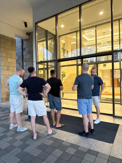 This image depicts a group of five men standing outside a modern building with large glass doors. The man on the far left is wearing a light blue polo shirt and striped shorts, with his back to the camera. Next to him, a bald man in a black t-shirt and striped shorts is also facing away, with his hands on his hips. A third man, also bald, in a dark grey t-shirt and blue shorts, stands in the center, facing towards the building. To his right, a man in a navy blue polo shirt and striped shorts is conversing with him, also facing the building. The man on the far right is partially visible, wearing a navy blue t-shirt and striped shorts. The building's entrance features a grand staircase visible through the glass doors, illuminated by modern circular light fixtures. Two security cameras are mounted on the exterior wall near the entrance. The ground in front of the building is paved with grey and black square tiles, and a ribbed black mat is placed at the entrance. The architecture suggests a hotel or a similar commercial establishment. The lighting indicates it is either early morning or late evening. The location is identified as Razliv, Bulgaria.