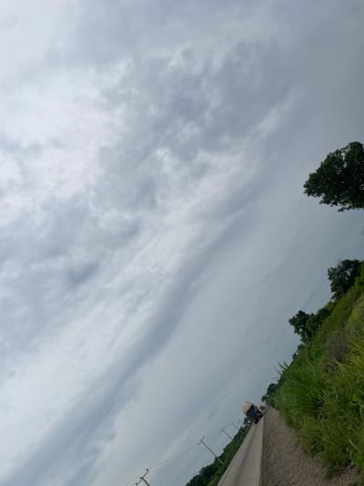 This media file captures a slightly tilted view of a road in Oduape, Nigeria, with lush green vegetation lining its right side. The sky is overcast with a mix of light and dark gray clouds, suggesting a cloudy day, possibly before or after rain.

A dark-colored truck is visible on the road, carrying a large, pale, round object on its back, possibly a large sack of produce or goods. The truck appears to be moving away from the viewer, down the road. Telephone poles with wires stretch along the side of the road, indicating the presence of infrastructure.

In the upper left portion of the image, the tops of trees are visible, their dark green foliage contrasting with the muted tones of the sky and road. The overall scene evokes a sense of rural or semi-rural life, with the road serving as a connection through the landscape. The mood is somewhat subdued due to the overcast weather. No visible text is present in the image.