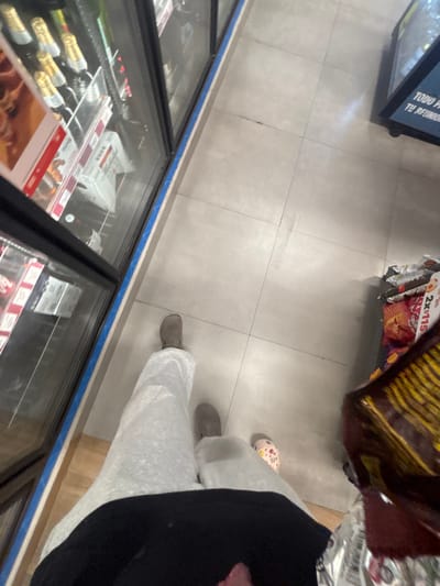 A downward shot inside a store, focusing on the tile floor and lower portions of refrigerated display cases. Bags of chips and other snacks are visible at the top. A person stands in the frame, wearing gray footwear and white pants, with a dark top partially obscuring the view. The display cases contain bottles and packaged items, with red promotional materials visible.