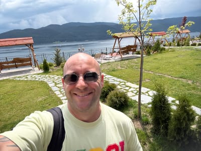 A bald Caucasian man with sunglasses and a light yellow t-shirt is taking a selfie in front of a body of water and a mountainous landscape. He is smiling at the camera. In the background, there are wooden swings with red roofs, a paved walkway, green grass, and trees. The scene appears to be a resort or park area near the Dospat Dam in Dospat, Bulgaria. The sky is cloudy.