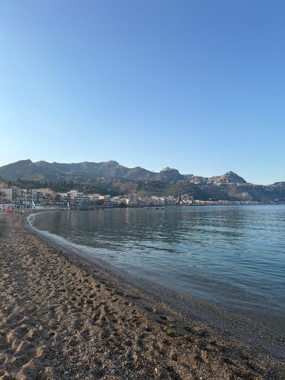 The image shows a coastal view in Chianchitta, Italy. A beach with dark sand or pebbles occupies the foreground, transitioning to the water. A town with buildings nestled on a mountainside extends along the coast. The sky is clear and blue. There are a few people and umbrellas visible on the beach.