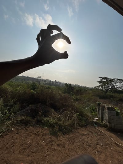 The image, reportedly captured in Debasco, Nigeria, displays an outdoor scene under a bright, partly cloudy sky. In the upper left quadrant, a silhouetted human left hand is positioned to partially frame and obscure the sun, which appears as a radiant disk with prominent light rays emanating from its circumference. The sky transitions from a light blue directly around the sun to a deeper blue further away, containing scattered white cumulus clouds.
Below the horizon line established by the hand, the foreground consists of uneven, reddish-brown earth mixed with sparse, dry vegetation and scattered debris. The mid-ground is dominated by an expanse of dense, uncultivated vegetation including various grasses, shrubs, and some smaller trees. Visible in the lower right, a stack of grey concrete blocks is present on a slight incline, alongside what appears to be a white plastic bag and a light-colored hose or pipe running along the ground. Further into the background, a large tree with a wide canopy stands on the right, and a hazy landscape featuring indistinct buildings and more greenery extends towards the horizon, indicating a semi-urban or developing area. No other individuals are visible in the scene.