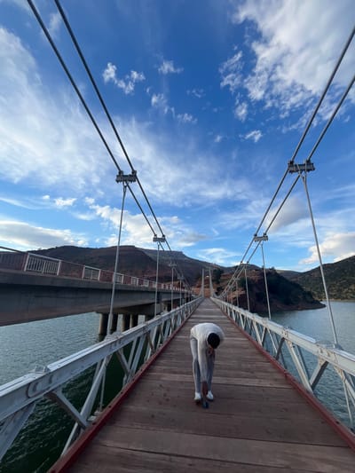 A man sits on a wooden suspension bridge in Ait Mazigh, Morocco. He wears light-colored clothing. The bridge spans a body of water and is surrounded by mountains. The sky is blue with scattered clouds. Steel cables and metal supports form the bridge's structure.
